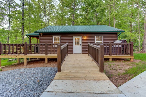 Log cabin with green metal roof, wooden deck, and railed walkway surrounded by tall trees