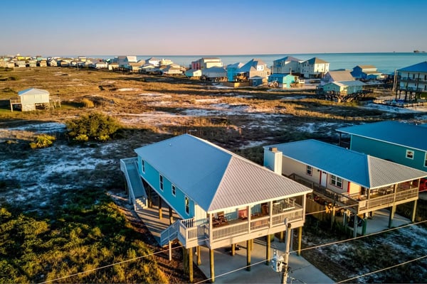 Elevated beach houses with blue roofs and wooden decks overlooking marshland and calm bay water, with more colorful houses lining the distant shoreline.