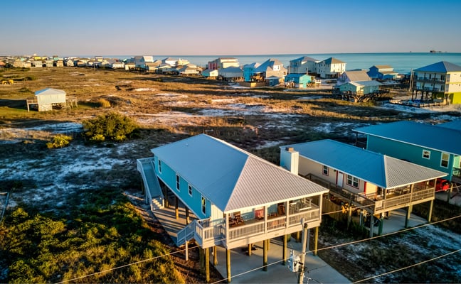 Elevated coastal homes with blue metal roofs on stilts overlooking marsh and bay, with additional houses lining the waterfront in distance